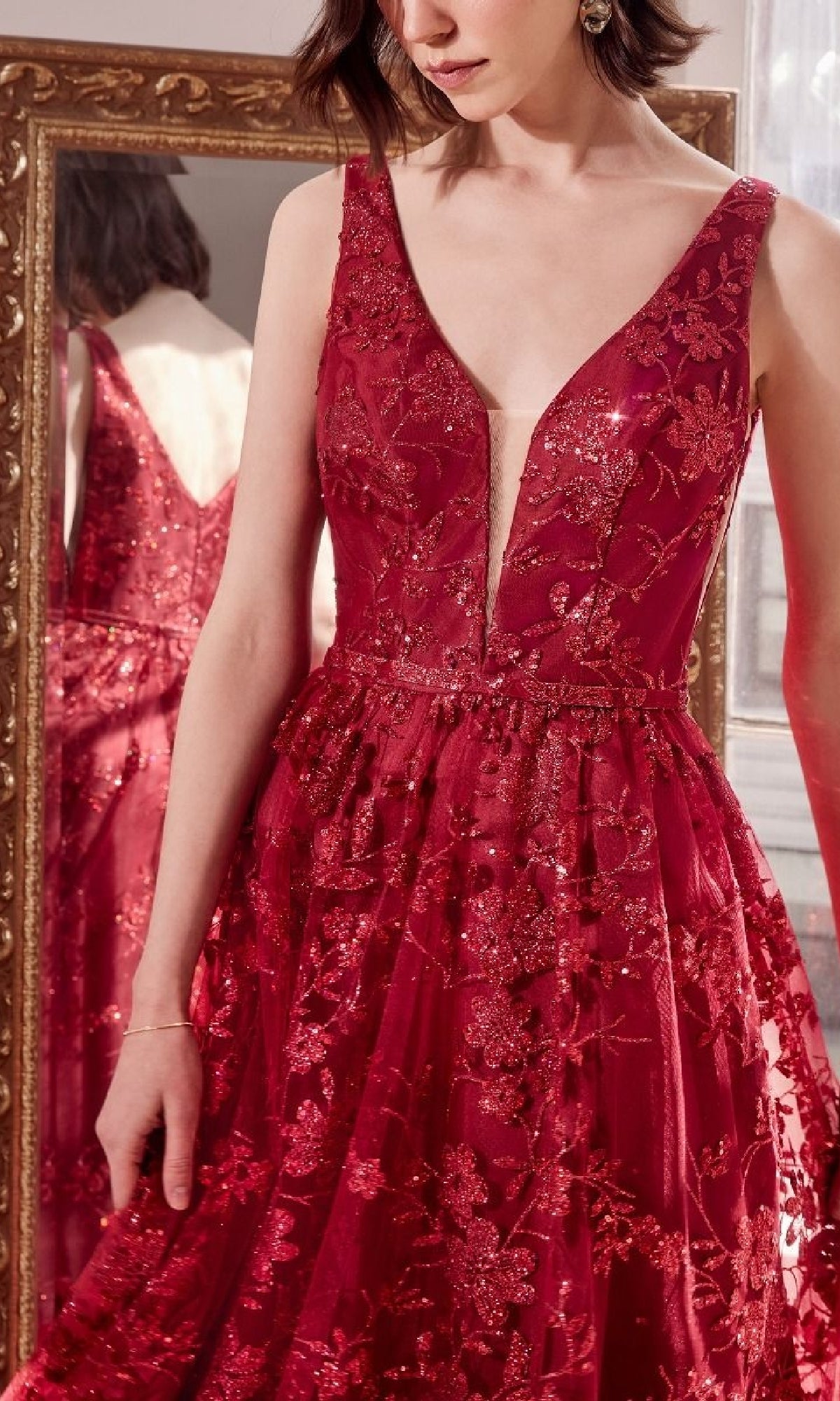 A woman stands in front of a mirror wearing the Embroidered-Lace Long Prom Dress: Amoris T606, a sleeveless gown featuring a deep V-neck, floral embroidery, and sparkling sequins.